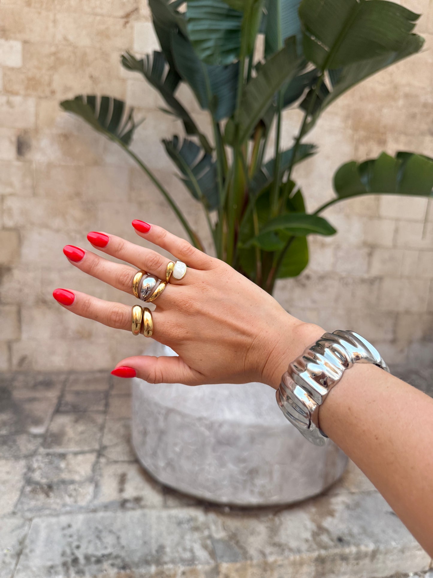 Hand with red nail polish wearing multiple rings and bracelets against a stone wall and plant background
