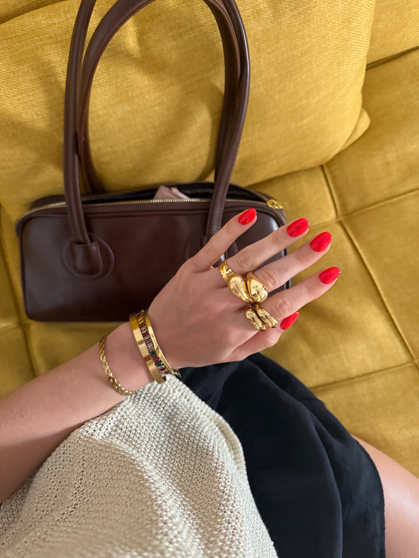 Hand with red nails and gold jewelry holding a brown handbag on a yellow couch.