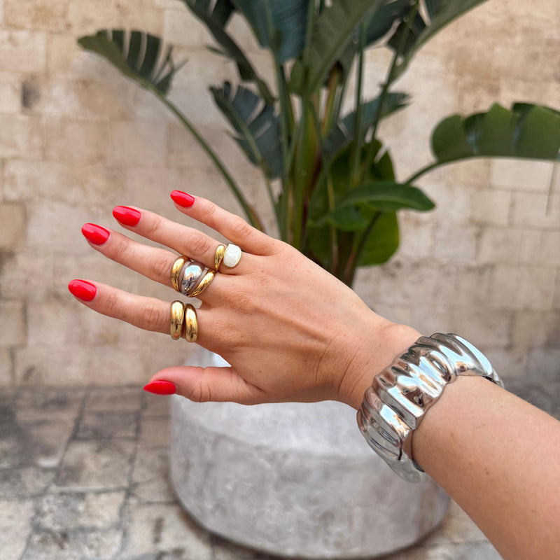 Hand with red nail polish wearing multiple rings and bracelets against a stone wall and plant background