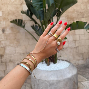 Hand with multiple gold rings and bracelets in front of a potted plant.