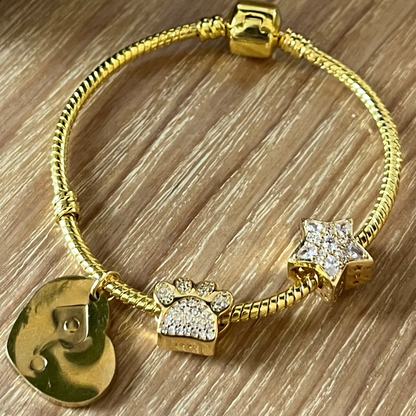 Gold bracelet with charms on a wooden surface next to a candle and mug.