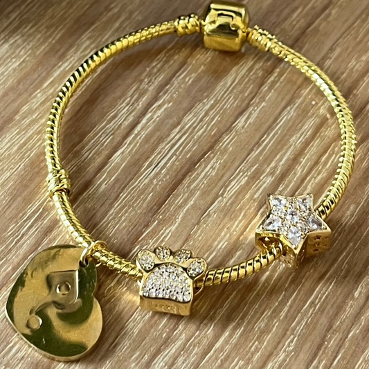 Gold bracelet with charms on a wooden surface next to a candle and mug.